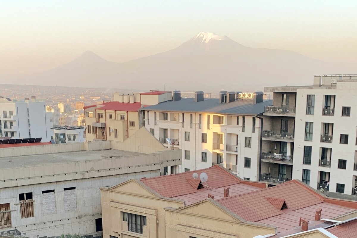 A view from a balcony captures the surrounding buildings against a backdrop of majestic Mount Ararat. The mountain's snow-capped peak is visible, with soft golden light illuminating part of the nearby architecture, evoking a serene urban landscape.
