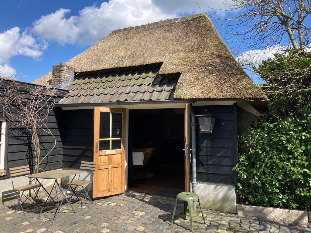 The entrance to a charming cottage features a thatched roof and wooden doors. A small outdoor seating area with a metal table and chairs is situated on a cobblestone pathway. Lush greenery frames the entrance, adding to the inviting exterior.