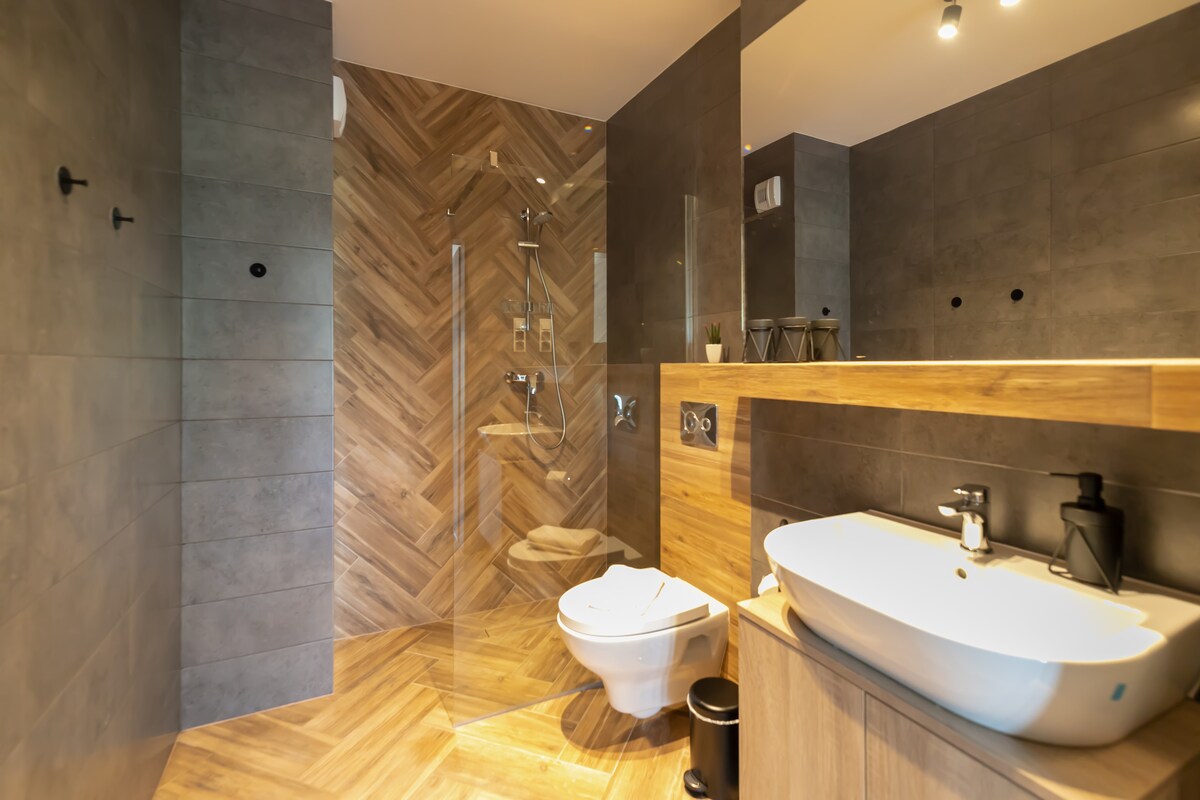 A modern bathroom features a walk-in shower with a sleek glass enclosure, complemented by wooden accents and contemporary fixtures. A stylish countertop basin is set atop natural wood cabinetry, while neutral tones and textured tiles create a balanced and calming environment.