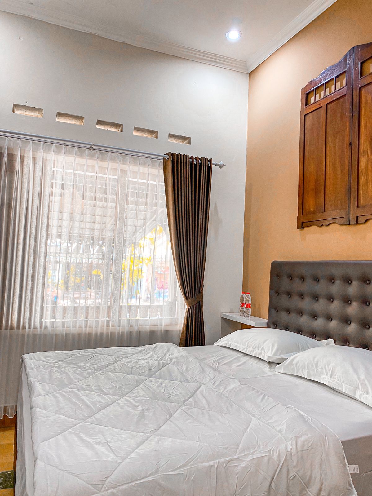 A modern guest room features a spacious bed with a gray upholstered headboard. Natural light filters through sheer curtains, illuminating the room's neutral tones. A wooden decorative panel enhances the wall, while bedside tables hold complimentary items.