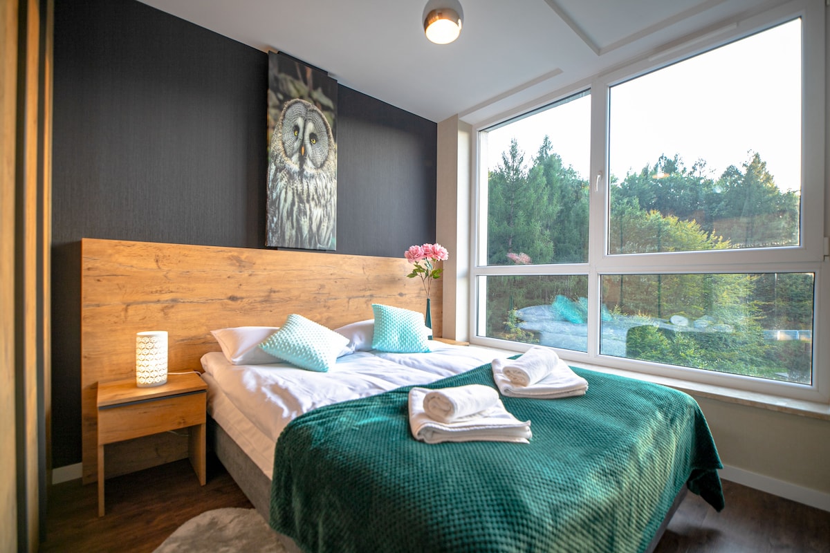 A brightly lit sleeping area features a spacious bed with a soft green blanket draped over it. Decorative pillows add a touch of color. A bedside table with a lantern is present. Large windows offer views of the surrounding greenery, enhancing the tranquil ambiance.