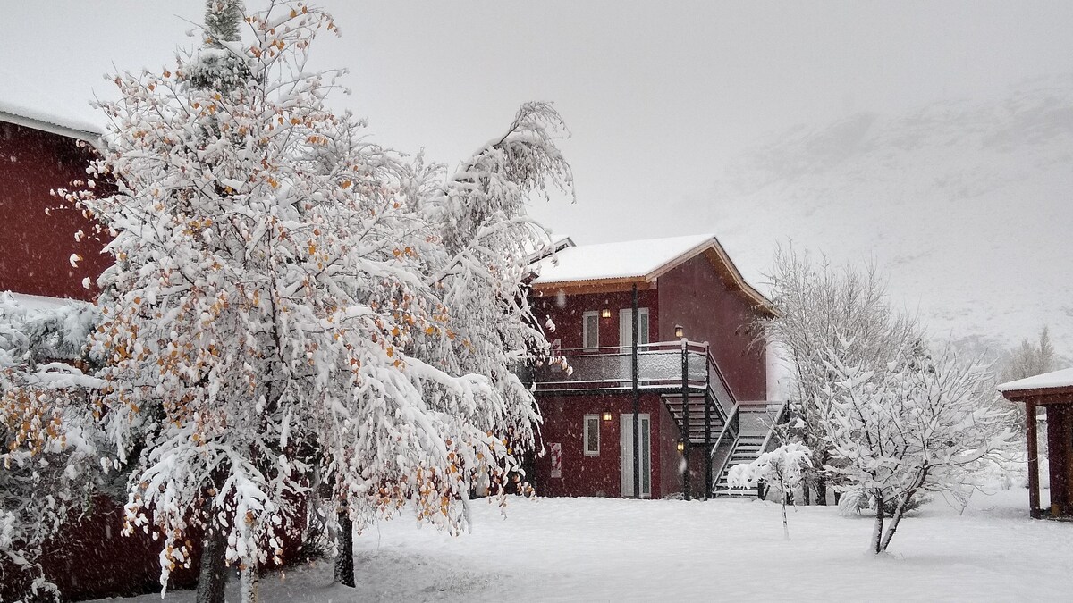 A snow-covered landscape features a two-story red building, surrounded by trees laden with snow. A staircase is visible leading to the upper level, and the scene conveys a serene winter atmosphere.