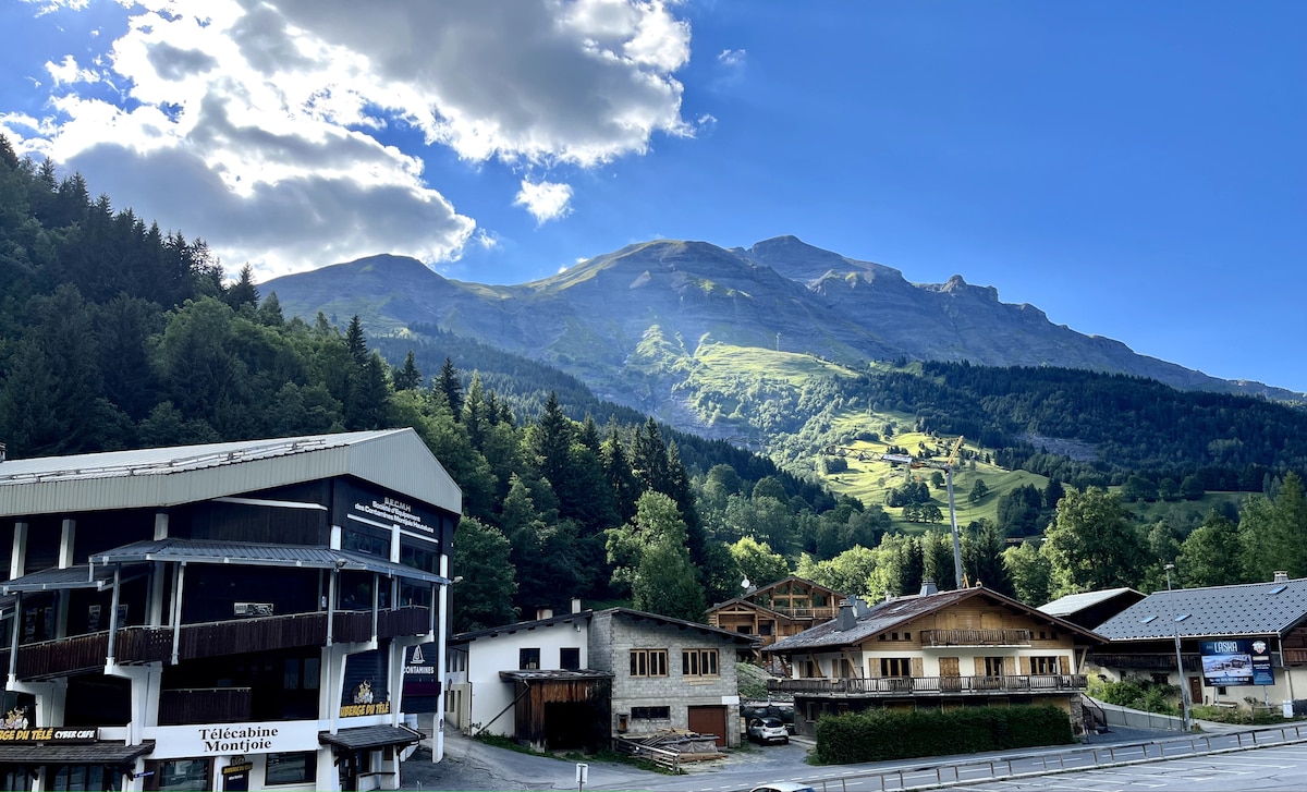 The image displays a scenic mountain landscape, showcasing green hills and towering peaks under a blue sky with scattered clouds. A mix of wooden buildings is positioned in the foreground, set among lush trees that complement the natural beauty of the location.