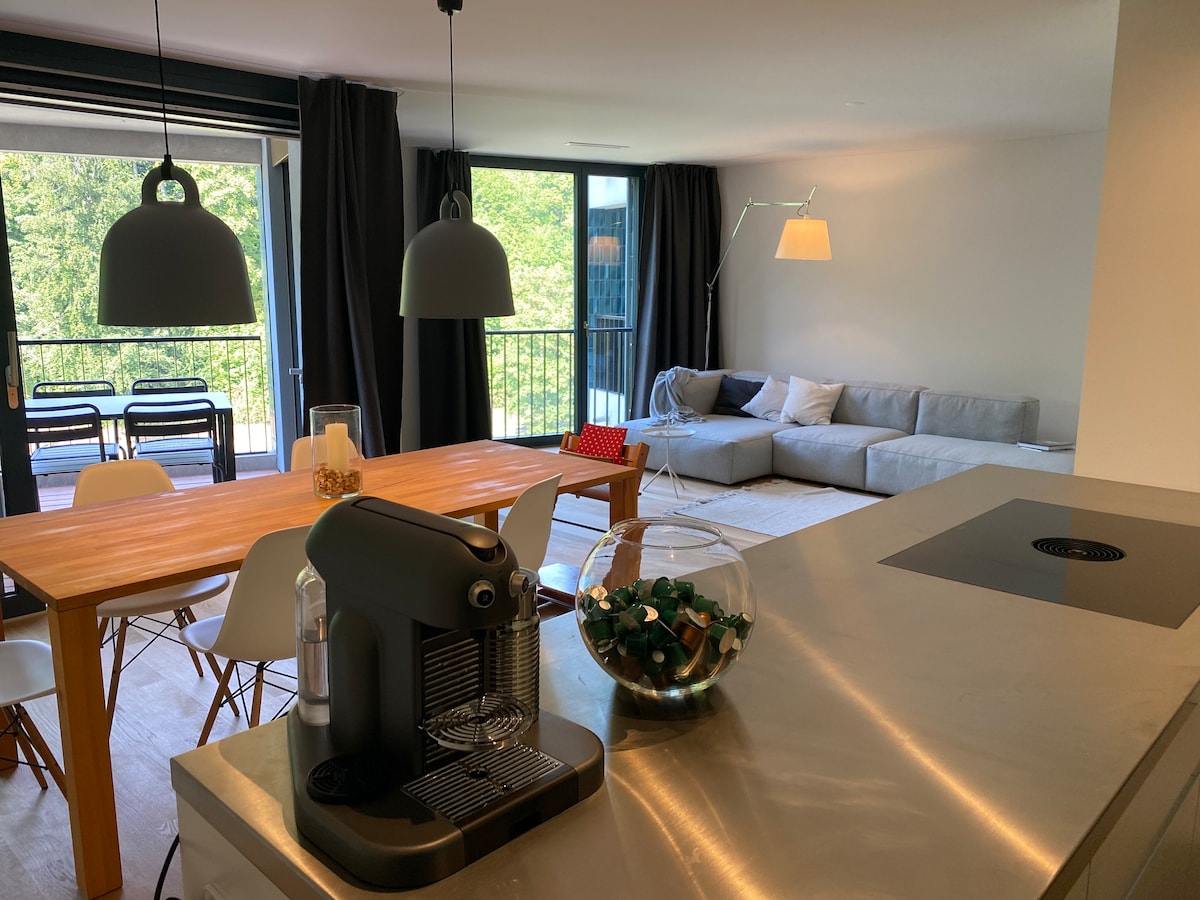 The open living area features a modern dining table, accompanied by stylish seating. A coffee machine is placed on a sleek countertop, while large windows offer views of the surrounding greenery. Soft lighting from pendant lamps enhances the space's inviting ambiance.