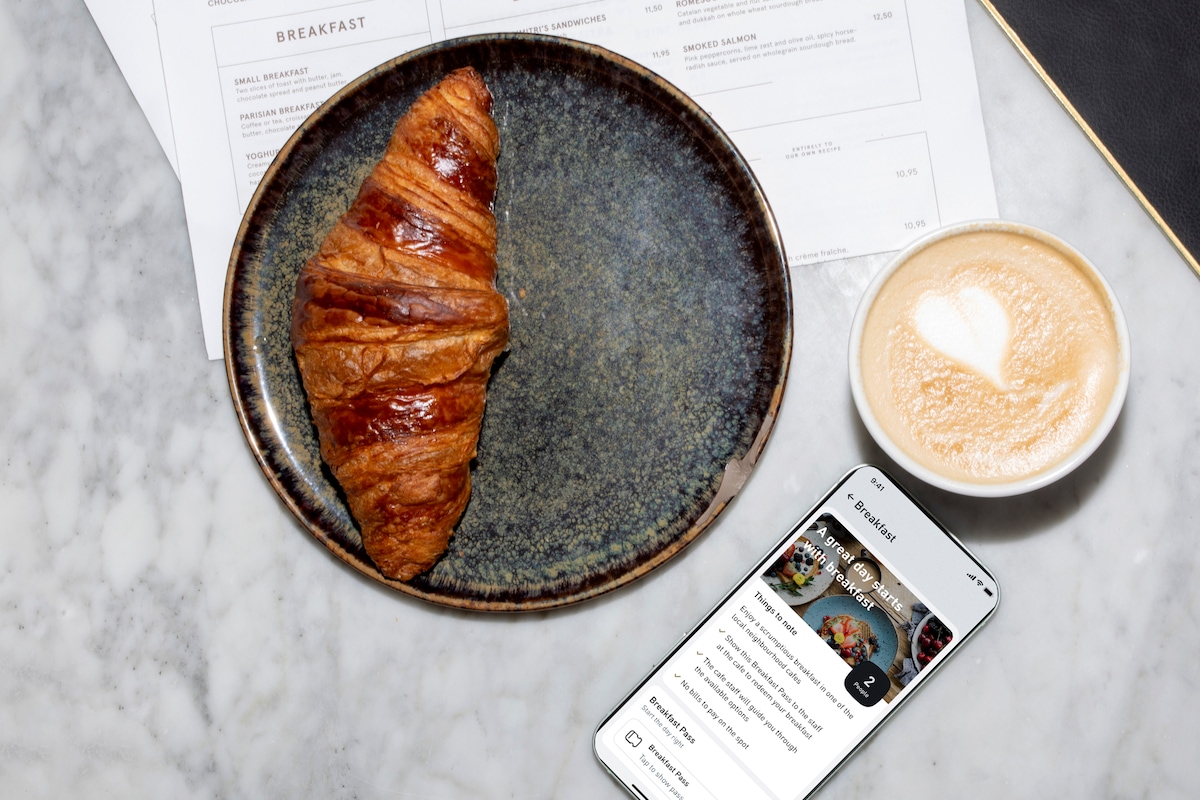 A croissant is presented on a dark, textured plate alongside a cup of cappuccino featuring a heart design in the foam. A mobile phone displaying a breakfast menu is placed on the marble surface, enhancing the breakfast setting.