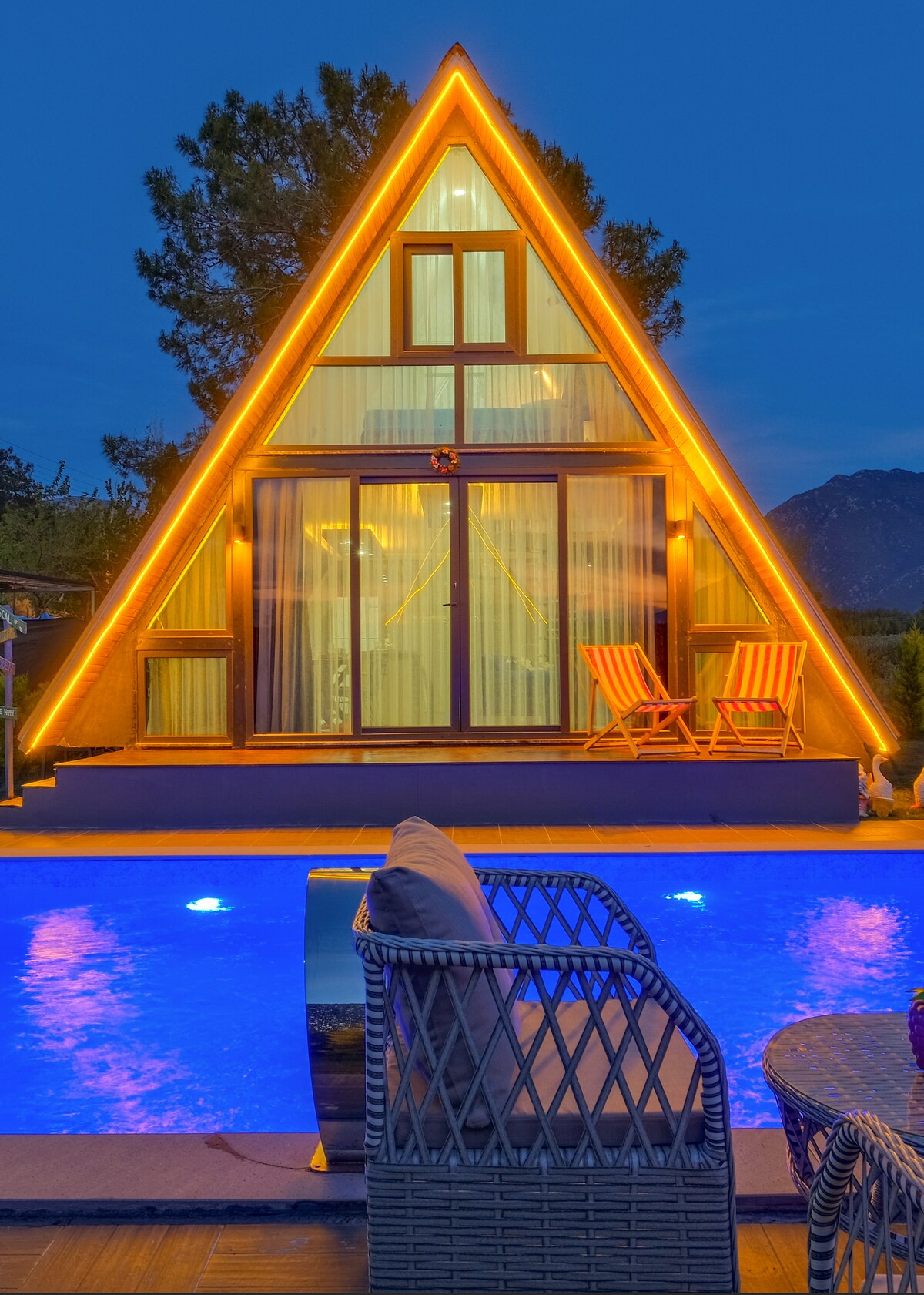 A modern triangular bungalow is highlighted by warm exterior lighting, standing beside a vibrant blue pool. Two striped chairs are positioned on the patio, offering a relaxing view of the serene surroundings.