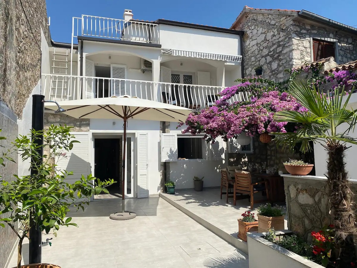 A spacious courtyard is enhanced by a large umbrella providing shade over a dining area. Lush greenery and vibrant purple flowers create an inviting atmosphere. The modern house facade features dual balconies, and the area is lined with potted plants, blending comfort and nature.