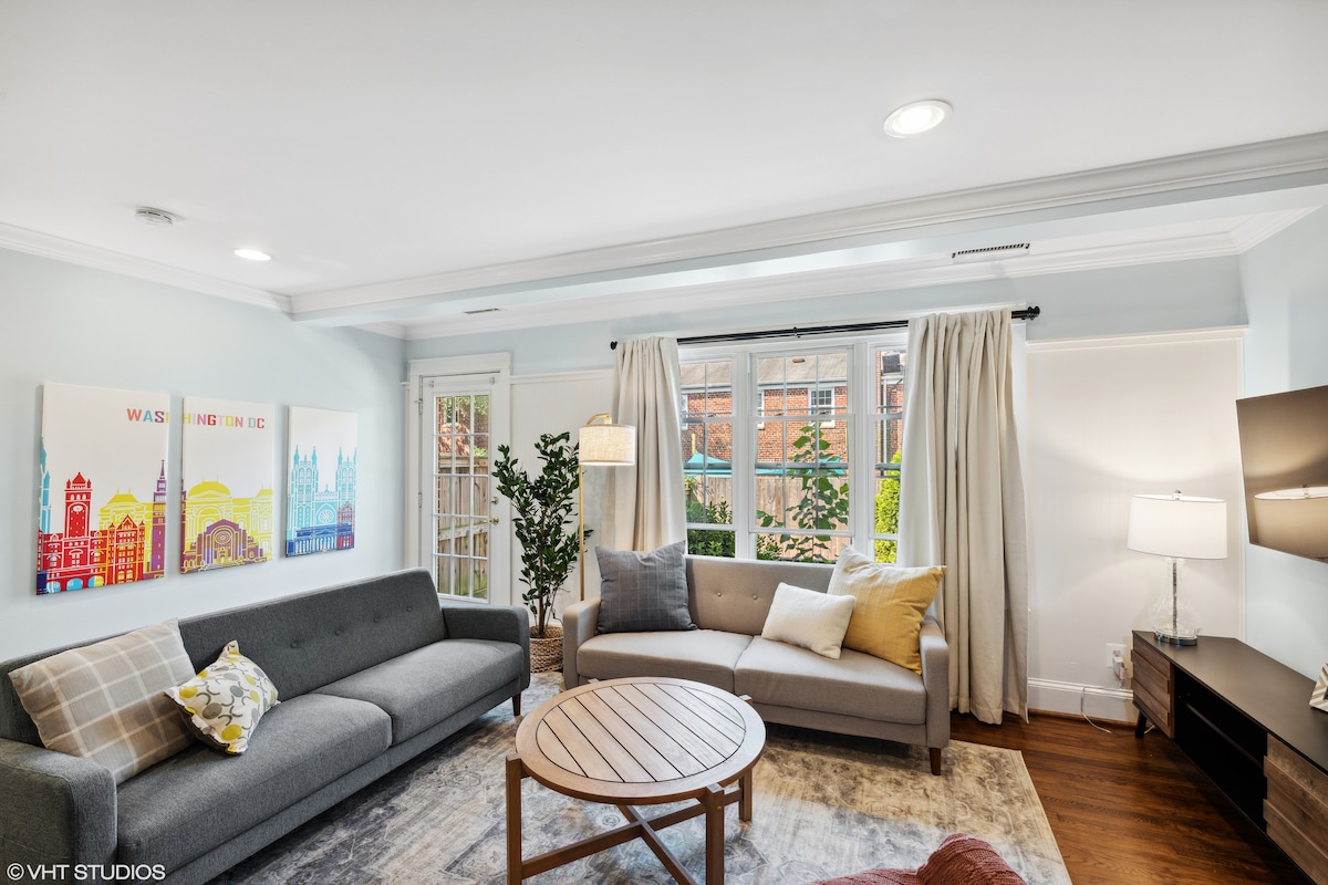 The living room features a spacious gray sofa and a round coffee table, complemented by a neutral area rug. Large windows with light curtains allow natural light to brighten the space, revealing artwork depicting Washington, D.C. in vibrant colors on the wall.