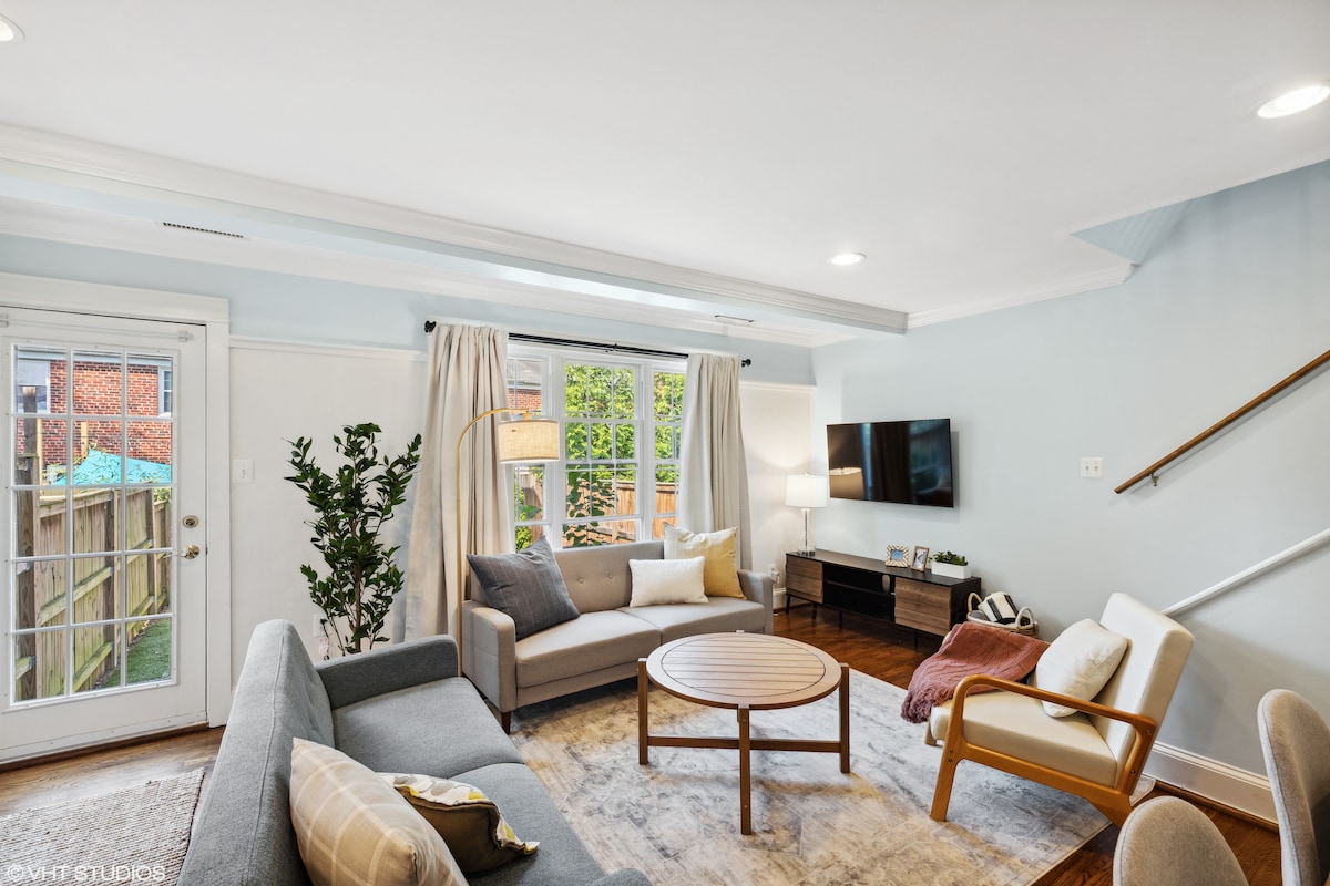 An inviting living area features a blend of comfortable seating, including a gray sofa and an armchair, arranged around a circular wooden coffee table. Natural light filters through large windows with sheer curtains, illuminating a soft area rug that complements the space.