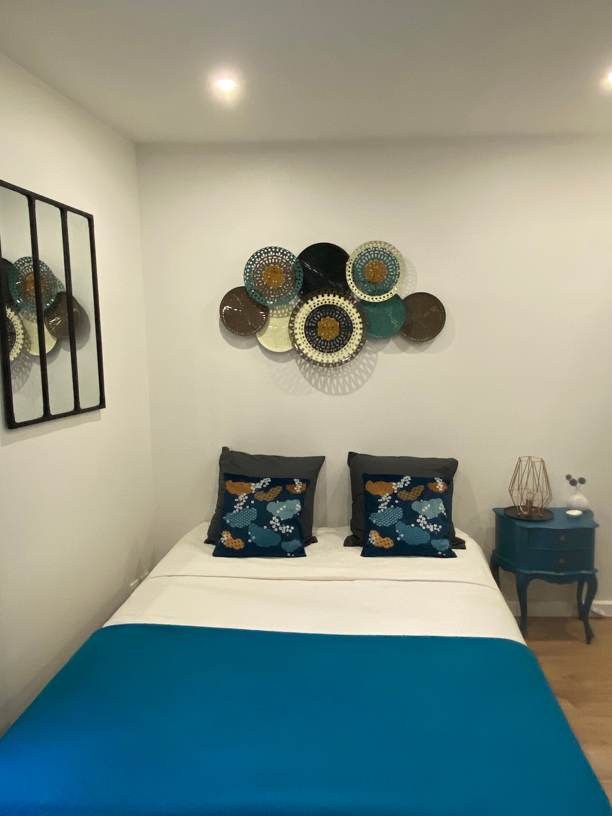 A compact sleeping area is furnished with a double bed, adorned with a white bedspread and a vibrant blue throw. Decorative pillows in blue and contrasting colors accent the bed. Wall decor features an arrangement of colorful plates above. A small side table complements the decor.