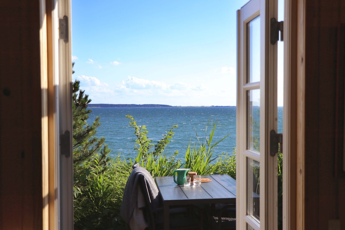Double doors open to reveal a serene view of Kalundborg Fjord, framed by lush greenery. A small table set for two sits on a deck, offering an inviting spot to enjoy the scenic water vista, while soft waves are faintly visible on the horizon.