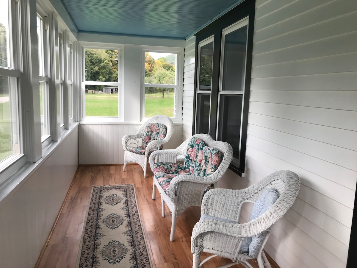 A sunlit porch features three white wicker chairs with floral cushions, positioned beside large windows. A softly patterned rug runs along the wooden floor, enhancing the space's natural light. The open view showcases greenery outside, inviting relaxation and comfort.