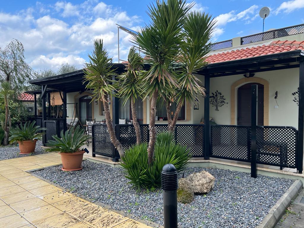 The exterior of the bungalow features a walkway lined with decorative stones and potted plants. Palm trees provide a tropical feel, while the porch showcases an inviting seating area beneath an overhang. The structure is complemented by a tiled roof and gentle landscaping.