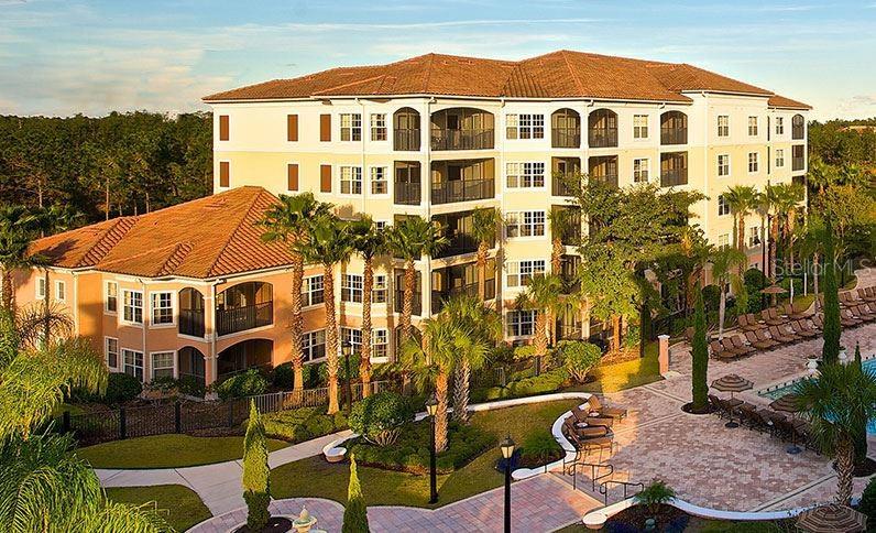 An expansive view of a multi-story condo complex surrounded by lush greenery and palm trees. The building features multiple balconies and large windows. The well-maintained landscaping includes manicured bushes and a pathway leading towards a resort-style area. A scenic backdrop enhances the peaceful ambiance.