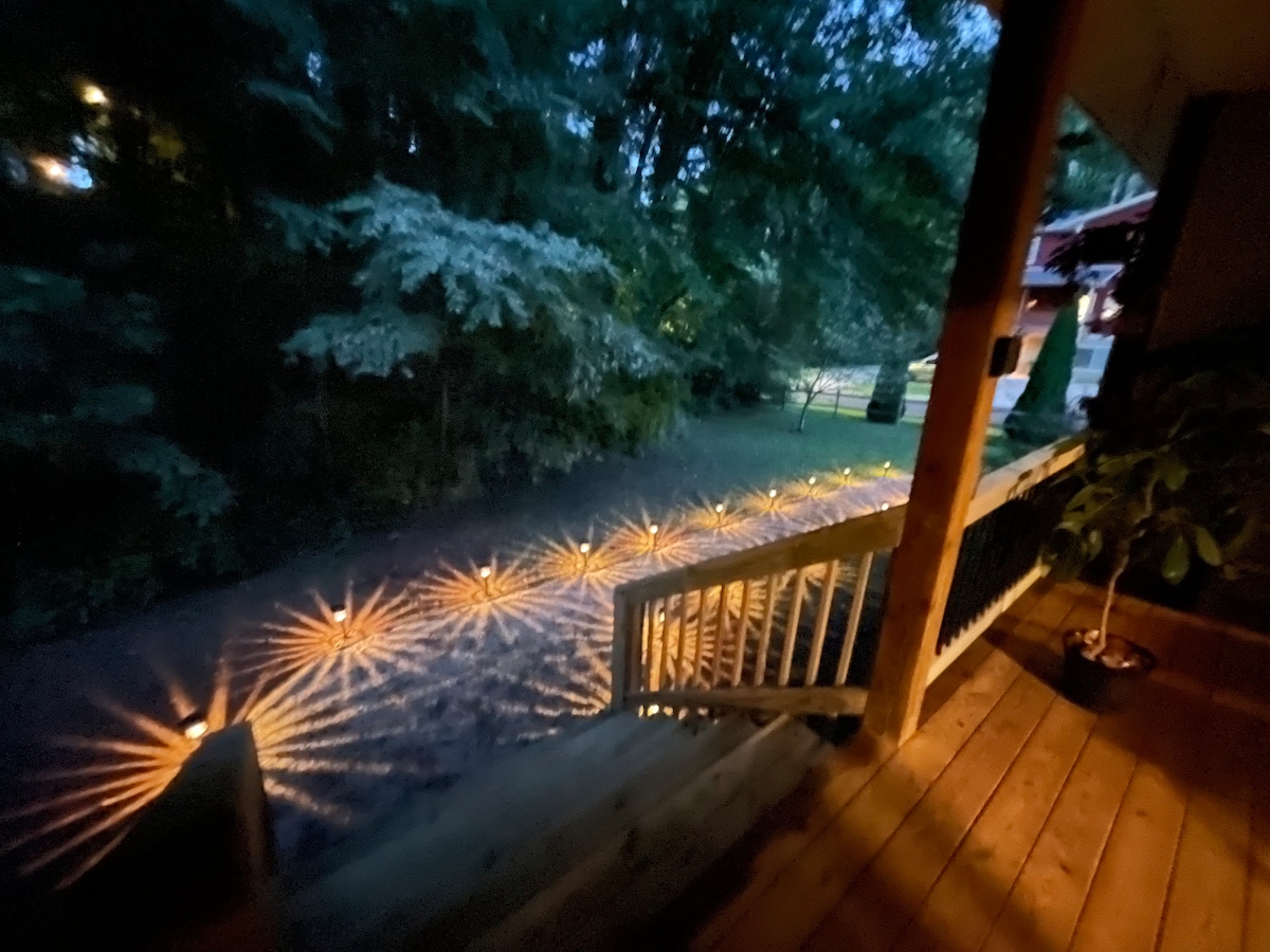 The entrance features a wooden deck with a railing, leading down toward a well-lit path. Decorative lights create a warm glow along the gravel path, surrounded by lush greenery. The peaceful ambiance is enhanced by the soft illumination from the surrounding lighting.