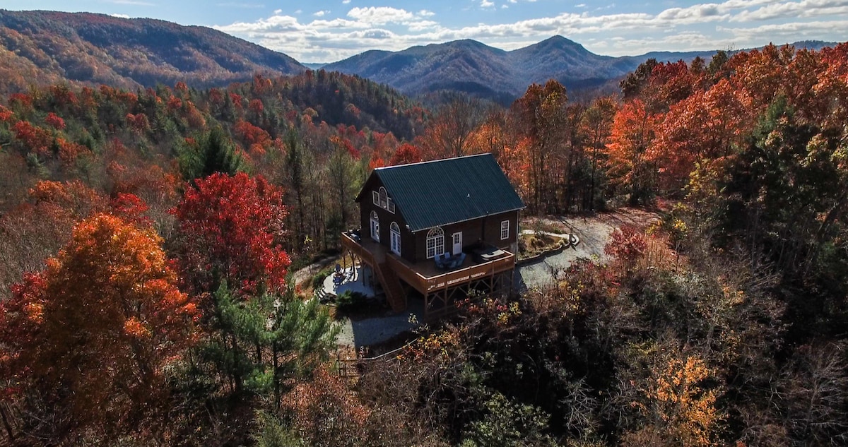 A three-story log cabin nestled in vibrant fall foliage, showcasing a blend of orange, red, and green leaves. The cabin features a welcoming deck and mountain views in the background, capturing the essence of a tranquil mountain retreat.