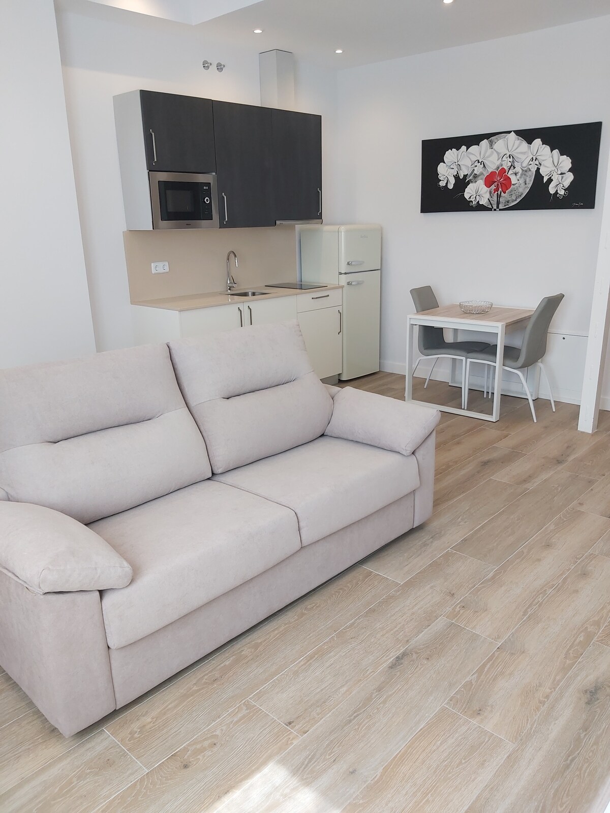 A modern living space features a light-colored sofa with textured cushions. An open kitchen area contains sleek cabinets, a microwave, and a refrigerator. A small dining table with four chairs is positioned nearby, creating a functional and welcoming atmosphere.