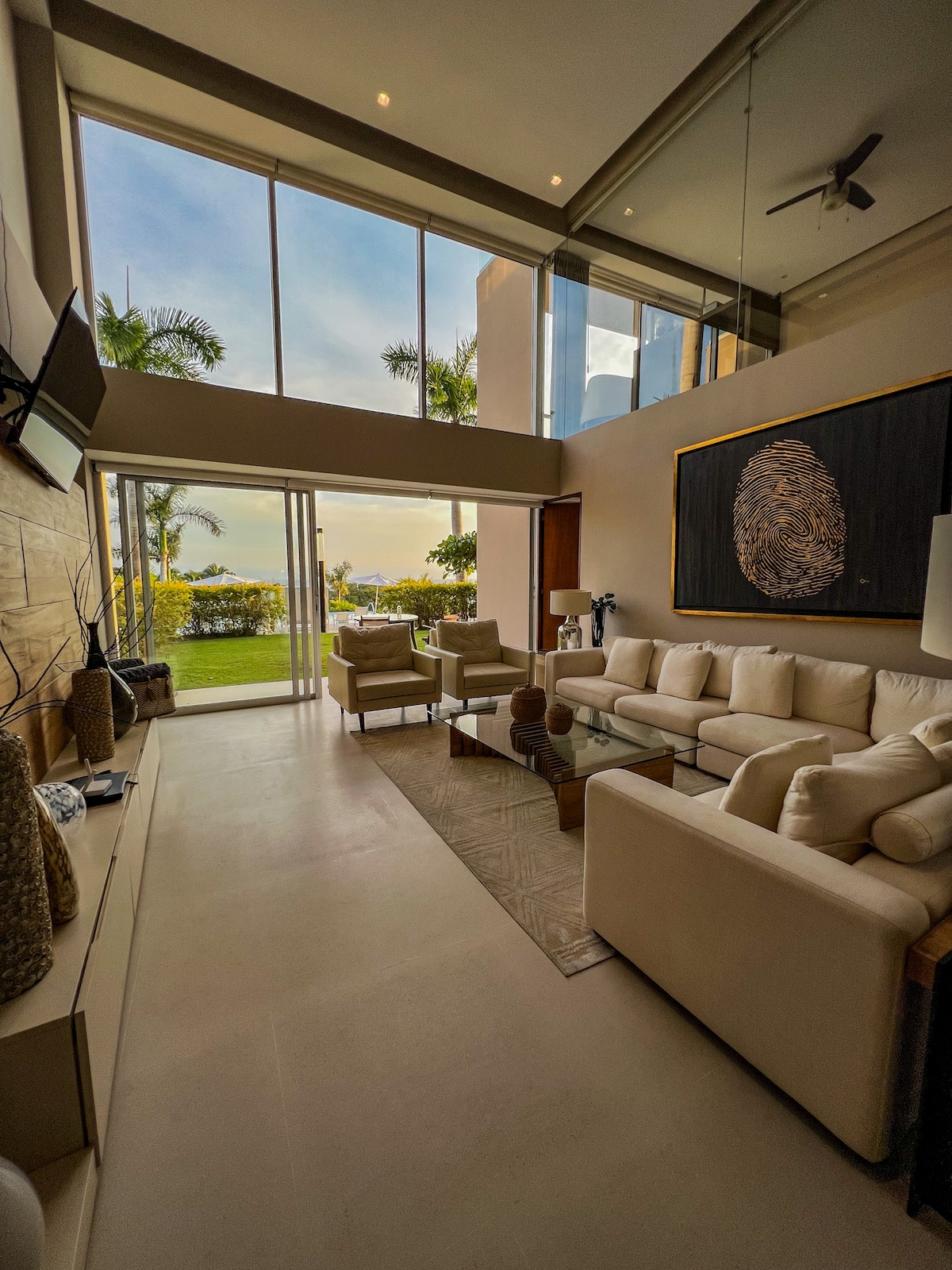 The spacious living area features large glass doors that open to a scenic view of the outdoors. Comfortable seating arrangements include light-colored sofas and chairs. Natural light floods the room, enhancing the modern design and subtle décor elements along the walls.