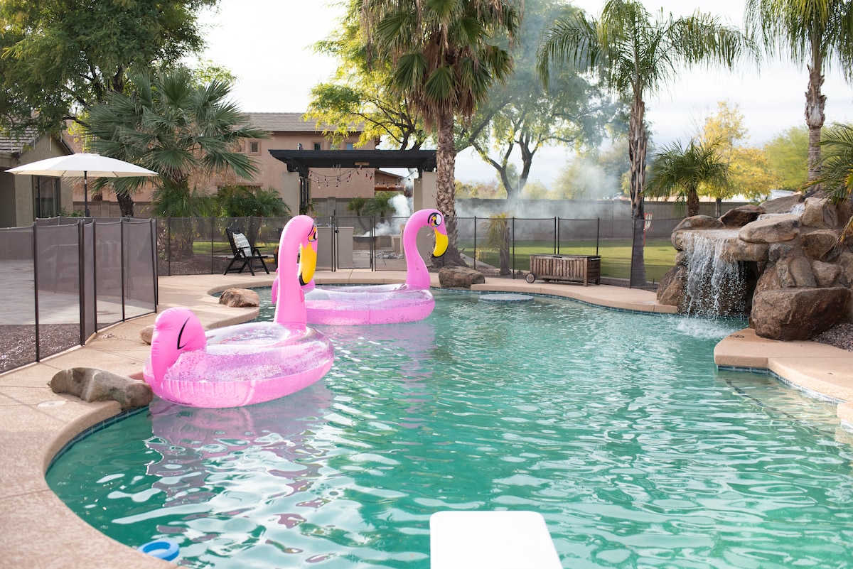 A private heated pool features pink flamingo inflatables floating on the water. Surrounding palm trees are visible against a clear sky, and a rocky waterfall adds a tranquil ambiance. A fenced area provides safety while lounge chairs and an umbrella are positioned nearby for relaxation.