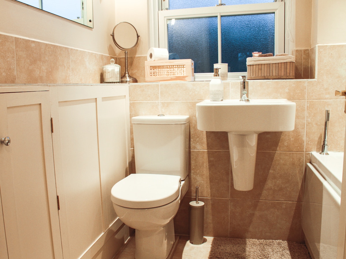 A well-organized bathroom features a clean, white sink and toilet, set against light beige tile walls. Storage solutions include a basket and a mirror. A bathtub is visible to one side, while natural light enters through a window, enhancing the overall cleanliness of the space.