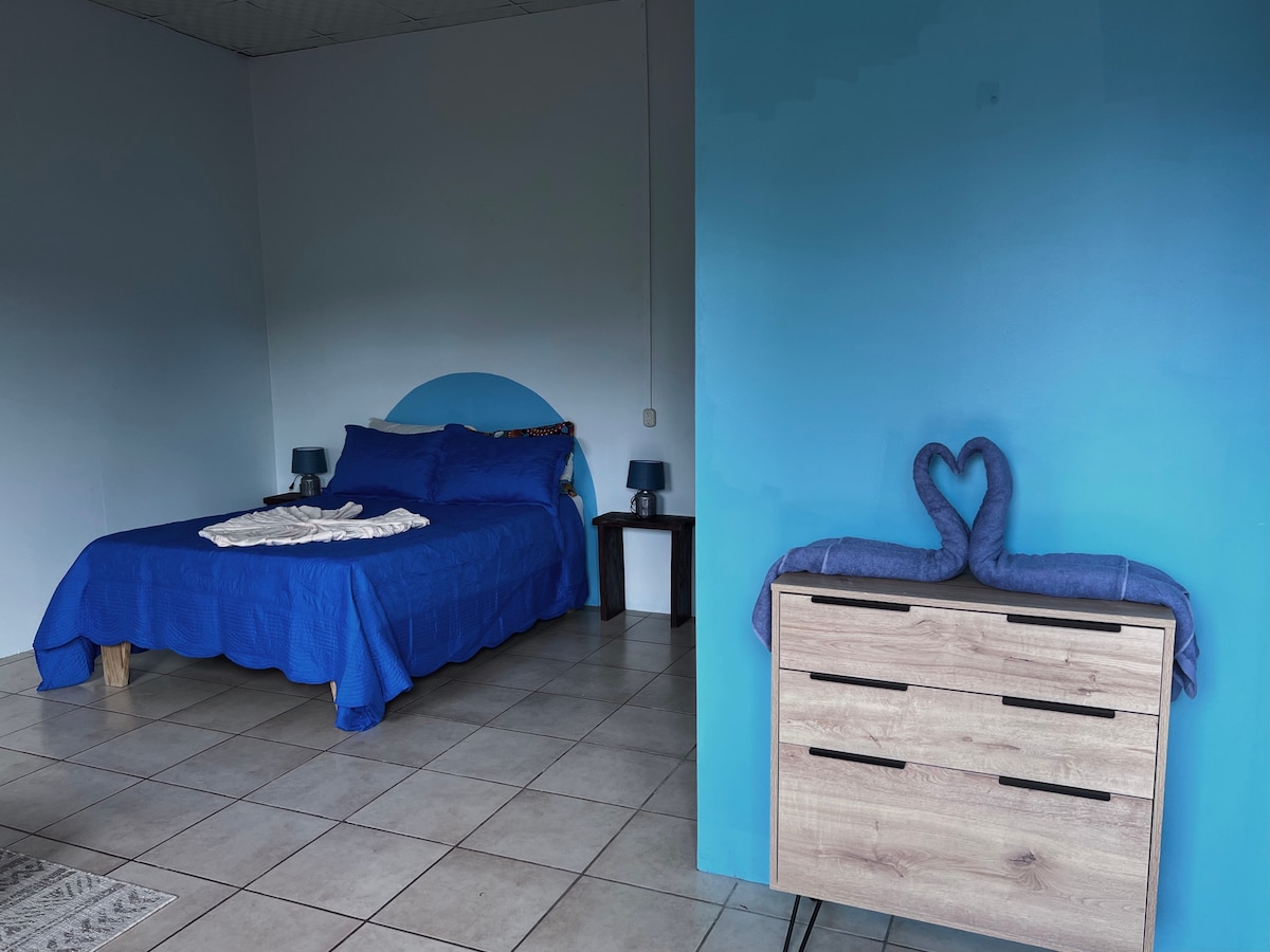 A spacious bedroom features a rich blue color scheme, with a large bed dressed in blue linens. A heart-shaped towel arrangement sits on a nearby dresser, which complements the room's decor. Soft lighting is provided by two bedside lamps.