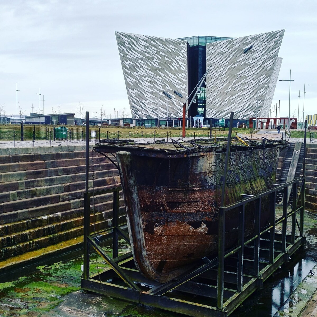 The image shows a rusted ship hull mounted on a framework, with a contrasting modern building featuring a unique angular design in the background. The area is surrounded by grass and walkways, illustrating a blend of historic and contemporary elements.