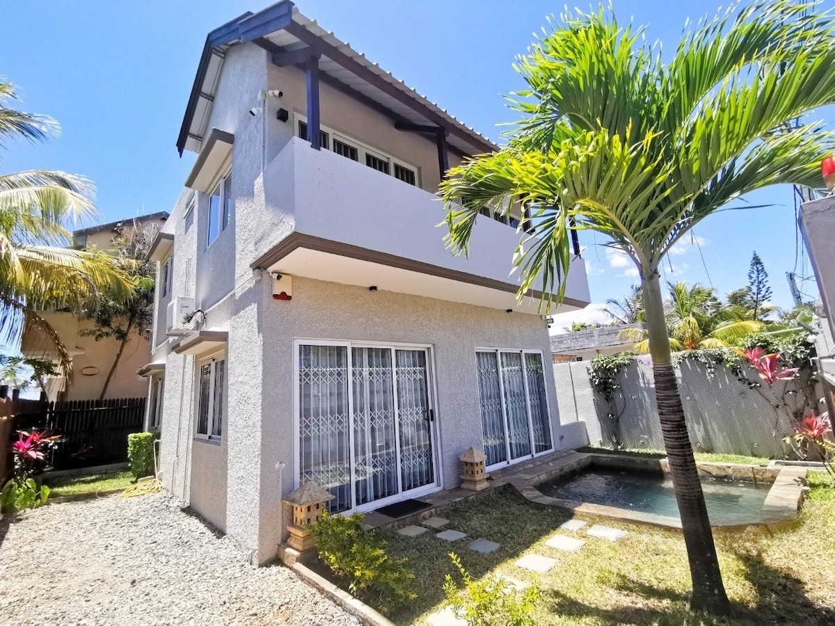 A two-story house is set in a landscaped garden, featuring sliding glass doors that provide access to a small plunge pool. Sunlight illuminates the exterior, which is complemented by a palm tree and decorative lanterns, enhancing the serene outdoor space.