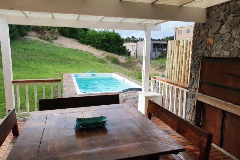 A covered outdoor seating area is outlined by wooden beams, featuring a table and benches crafted from dark wood. In the background, a private pool glistens under daylight, surrounded by a grassy landscape that gently slopes away.