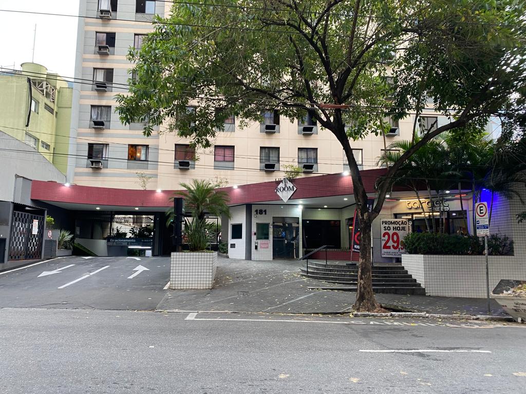 The building entrance is framed by a lush green canopy, highlighting a welcoming pathway. A decorative sign marks the entrance, and multiple vehicles are visible in the driveway. Windows adorned with air conditioning units suggest comfortable indoor environments.