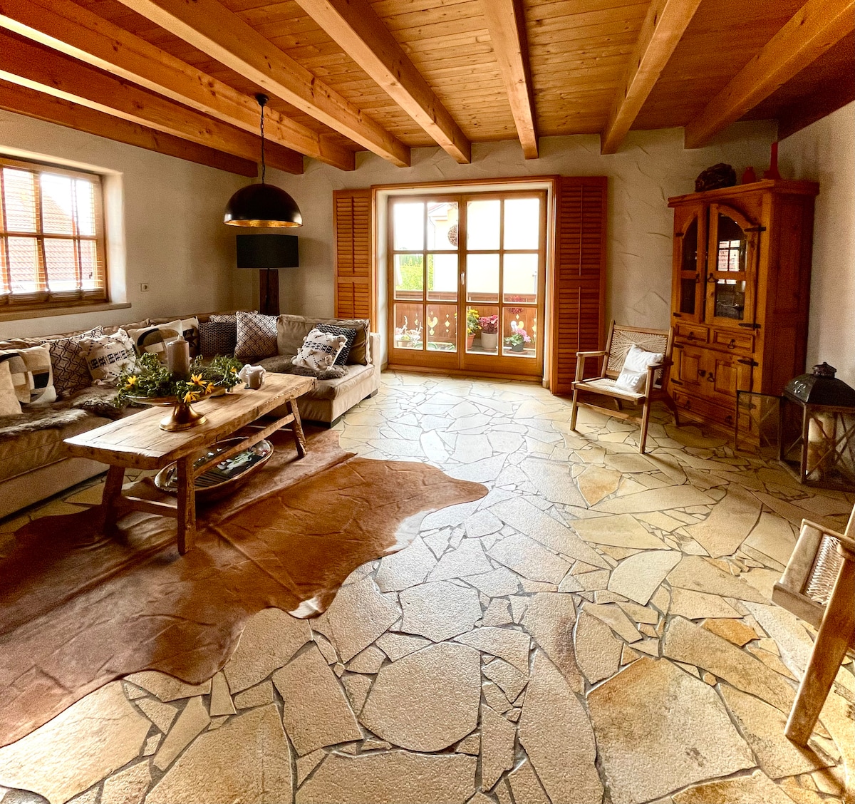 A spacious living area features a comfortable sectional sofa arranged around a wooden coffee table. Natural light filters through large glass doors that open to a balcony. The stone floor and wooden beams add warmth to the inviting space.