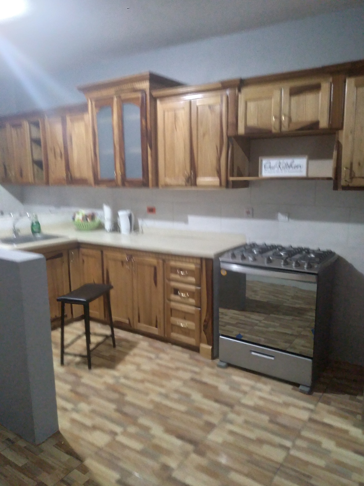 A functional kitchen is presented, featuring wooden cabinets with a polished finish and ample storage. A gas stove is positioned against the wall beside a countertop, while a tall stool adds seating. Natural light enhances the warm tones of the cabinetry.
