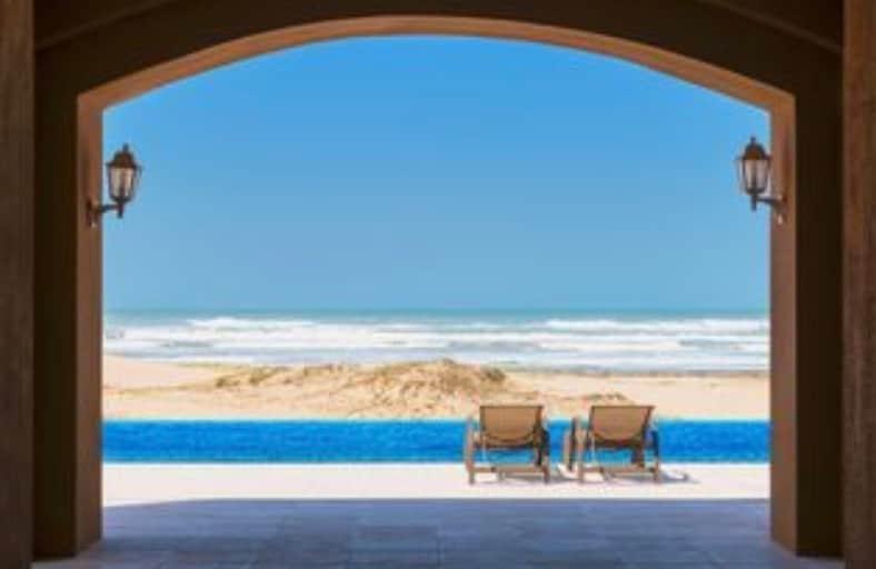A serene view showcases two lounge chairs positioned before a pristine beach. The ocean waves gently lap at the shore, while a blue infinity pool reflects the bright sky. Architectural arches frame the scene, enhancing the tranquil atmosphere.