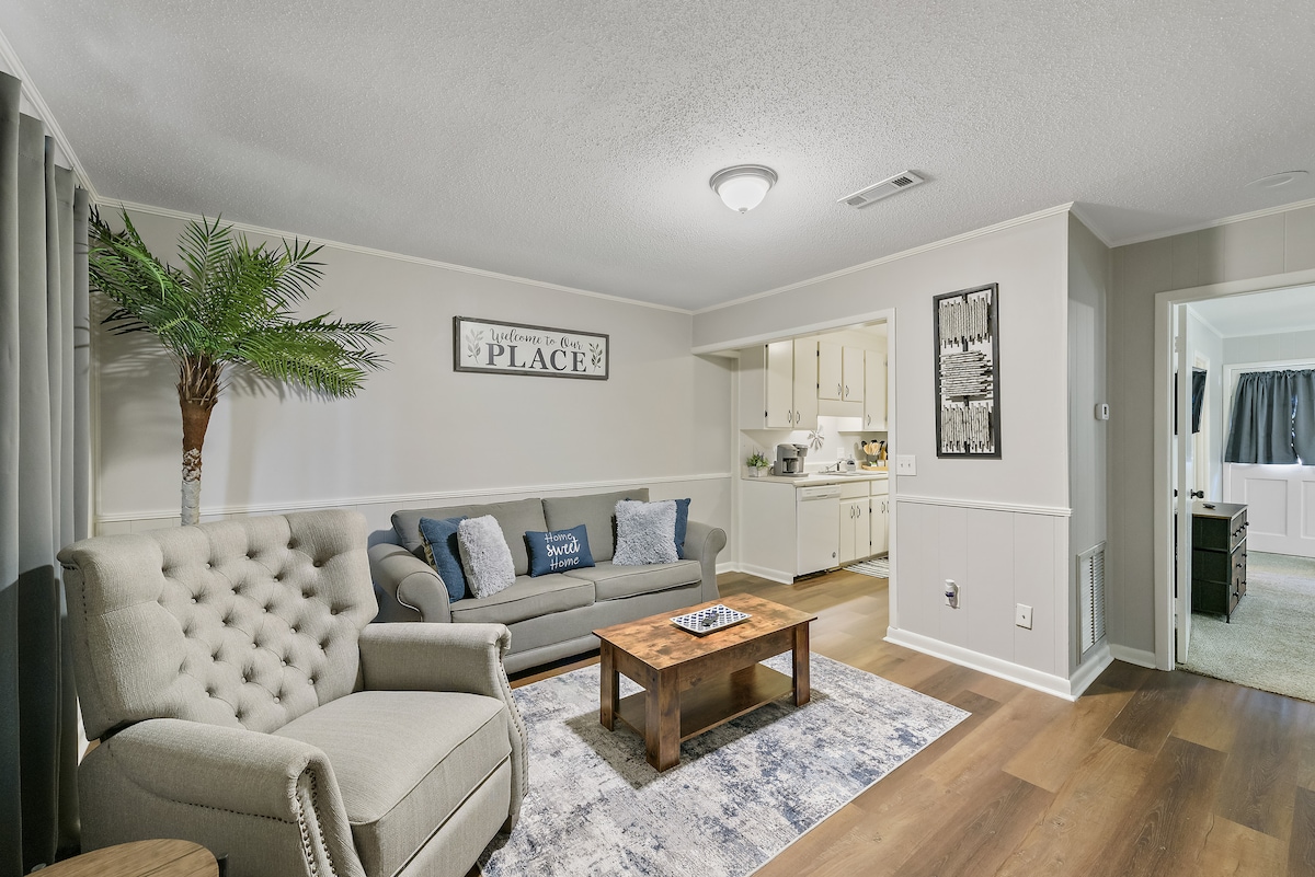 The living area showcases comfortable seating arrangements, including a tufted gray sofa and armchairs. Natural light fills the space, and a wooden coffee table sits atop a patterned rug. A view of the open kitchen is visible in the background, enhancing the spacious feel.