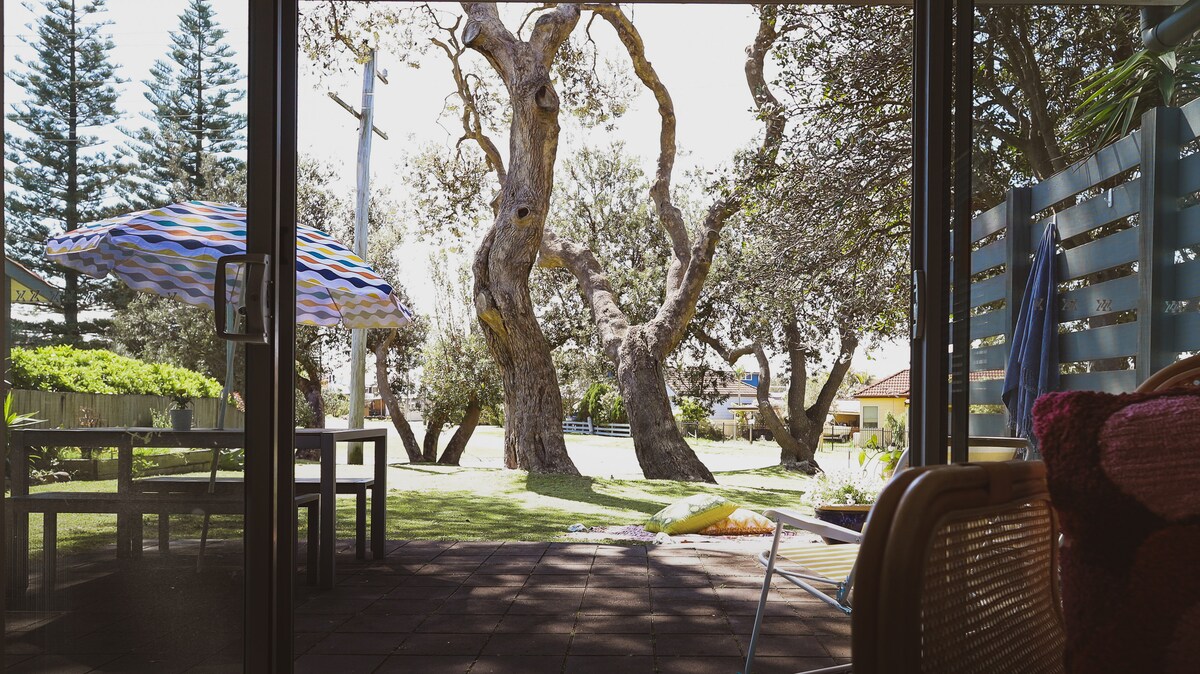 A view from inside the property showcases a spacious garden area framed by large, leafy trees. A colorful umbrella provides shade over an outdoor table, while the lawn extends towards the street, offering a peaceful retreat.