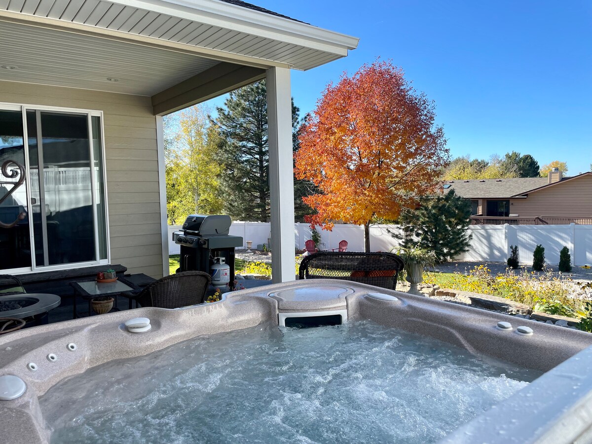 A hot tub filled with bubbling water is situated on a patio under a covered area. In the background, colorful trees display autumn foliage, while a grill and seating area are visible nearby. A fence borders the yard, enhancing the outdoor space.