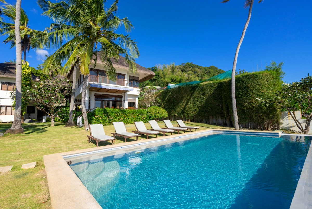 A spacious outdoor area features a glistening swimming pool surrounded by lounge chairs. Lush greenery and palm trees provide a natural backdrop, while a two-story building is visible in the distance against a clear blue sky.