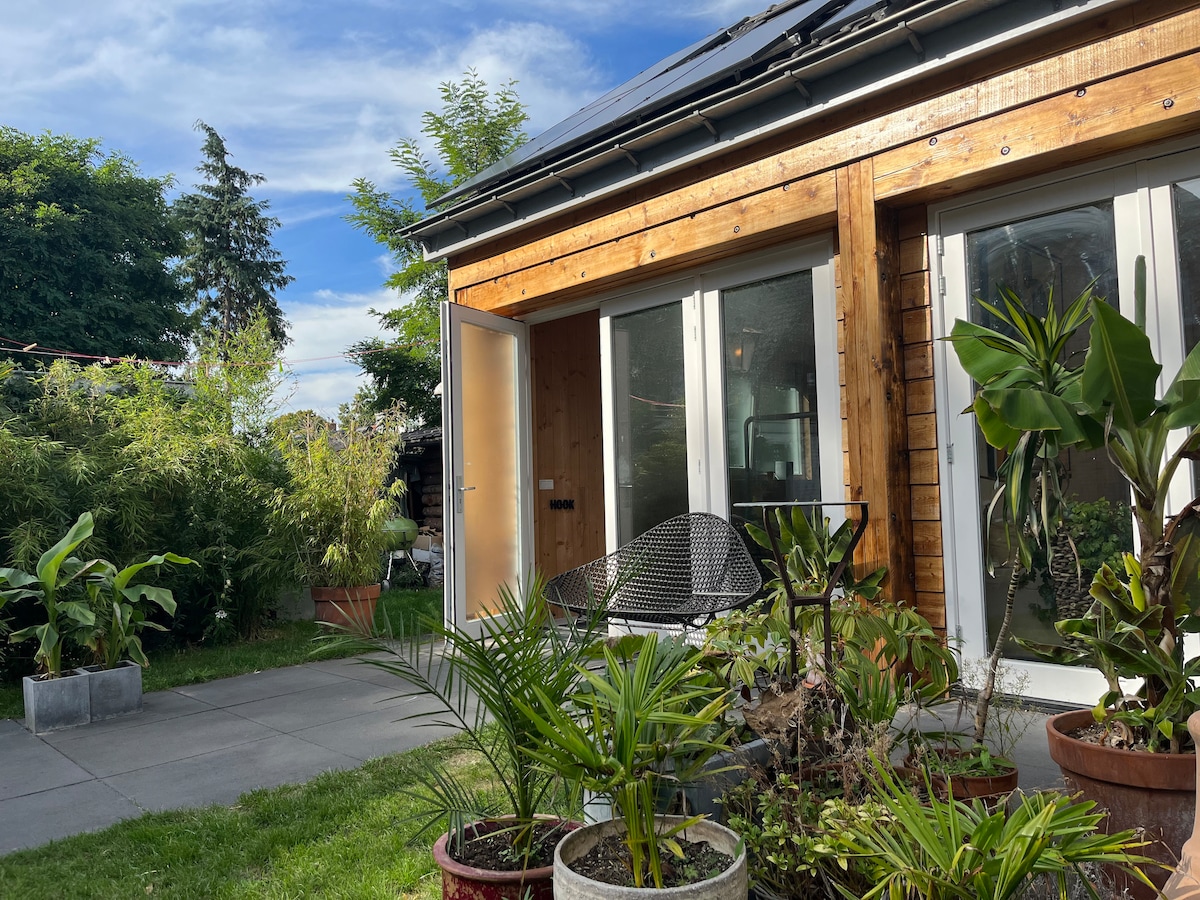 A wooden exterior is complemented by large glass doors, allowing natural light to fill the interior. Potted plants add a touch of greenery to the outdoor space, which features a well-maintained lawn and paved area. The sky is partly cloudy, enhancing the tranquil environment.