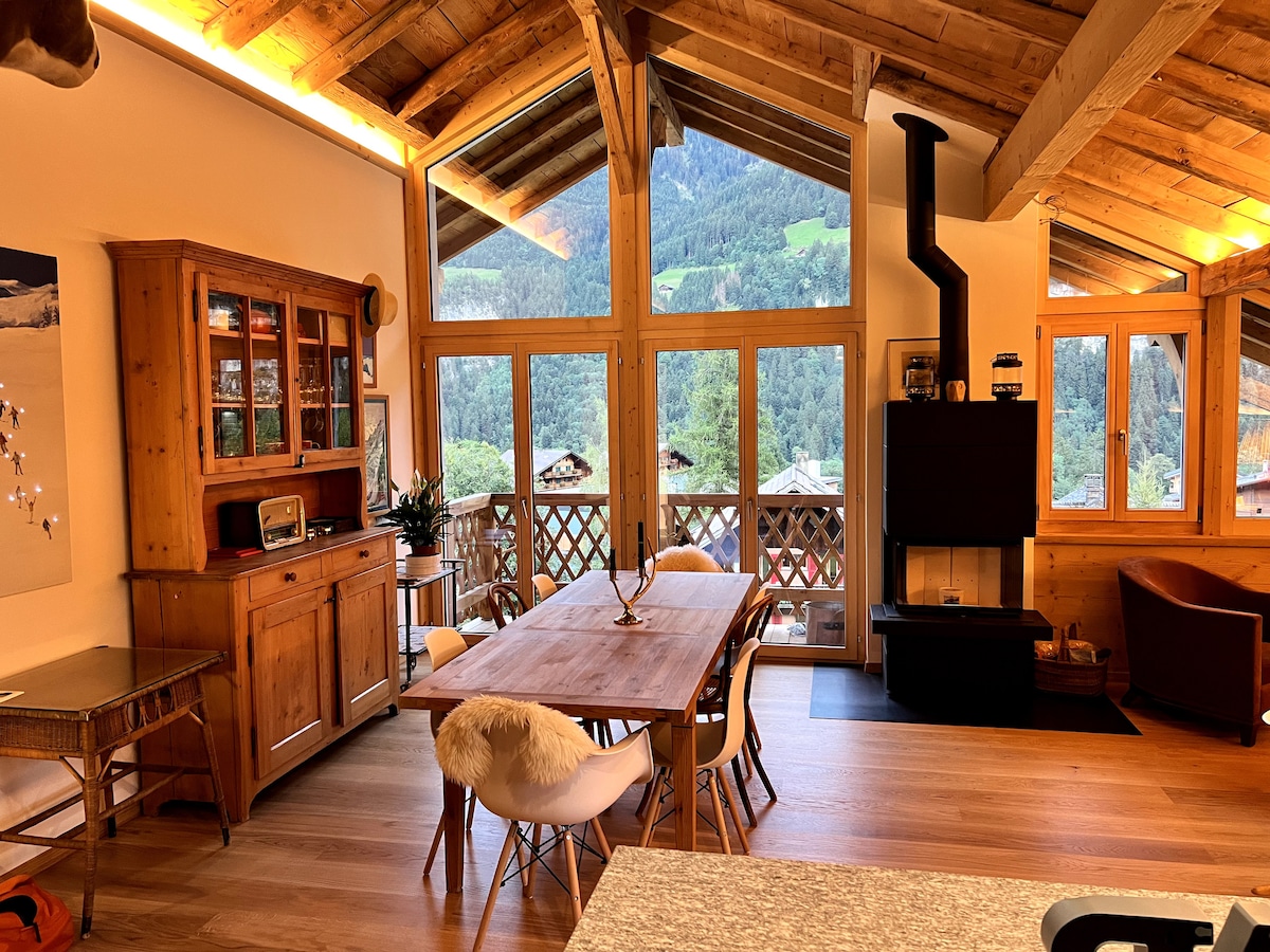 A spacious living area features large windows that frame mountain views. A wooden dining table is surrounded by chairs, while a cozy seating area showcases a modern fireplace. Natural light enhances the warm wood beams and the welcoming ambiance of the space.