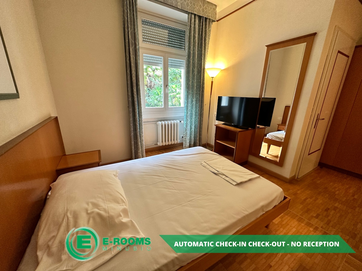A standard guest room features a spacious bed with crisp white linens, complemented by a bedside lamp and a flat-screen television. A large window allows natural light to enter, and a mirrored wardrobe adds depth to the space, creating a welcoming atmosphere.