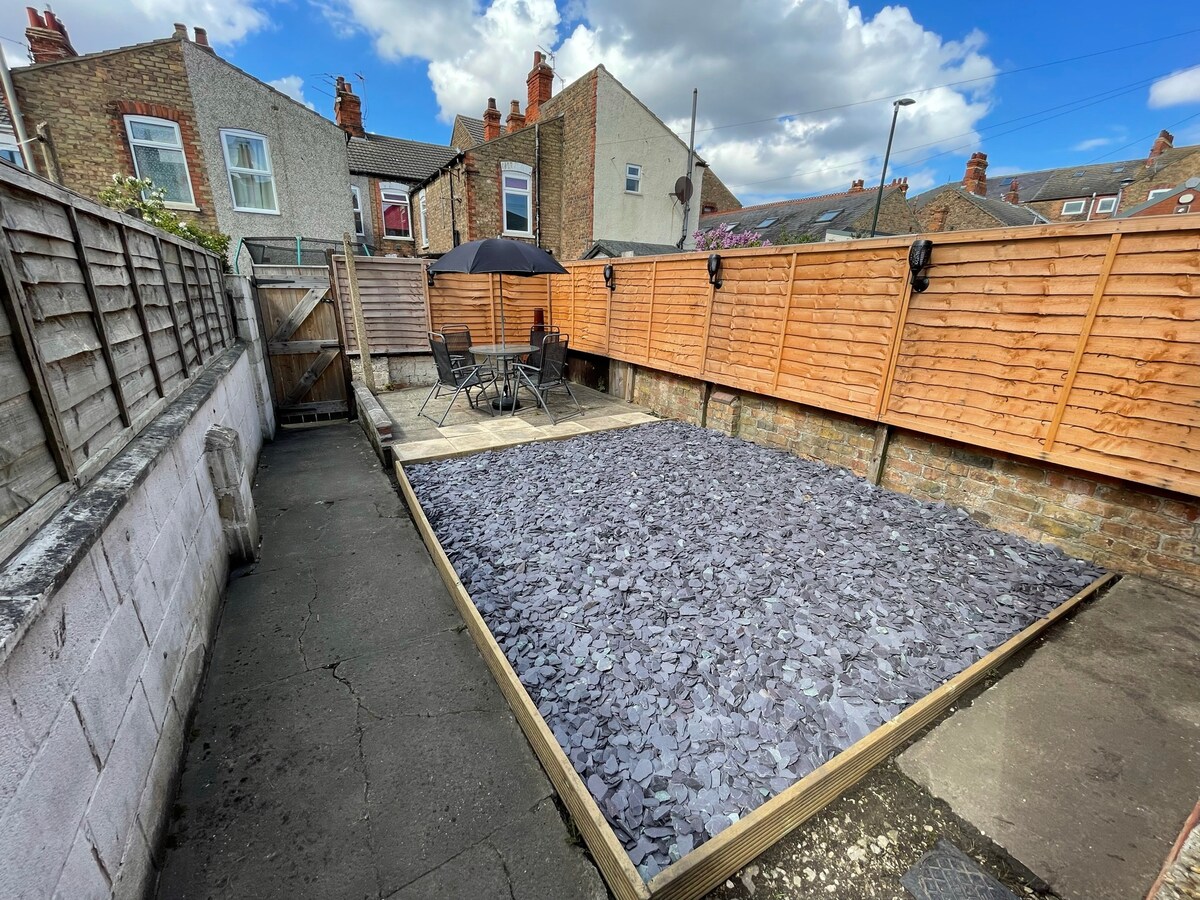 A private outdoor space is presented, featuring a wooden deck area surrounded by wooden fencing. A small table and four chairs are arranged beneath a black umbrella, providing a setting for relaxation. The ground is covered with slate stone, contributing to the overall tidy appearance.