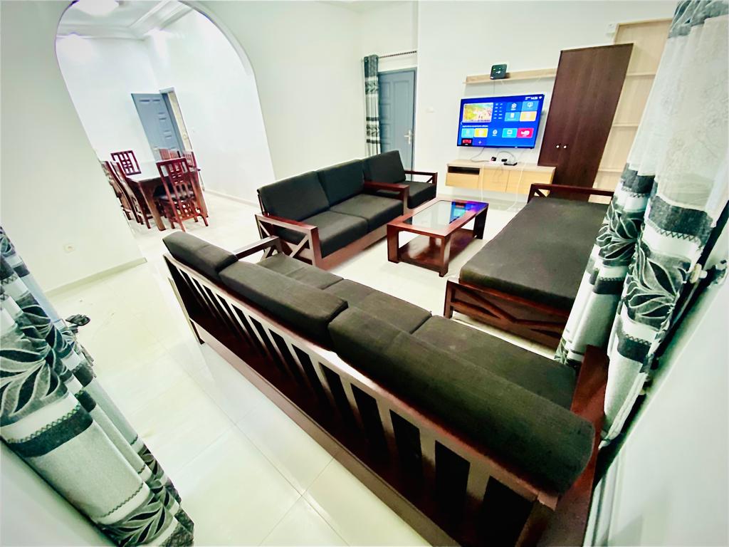 A spacious living area features a comfortable seating arrangement with dark wooden furniture and black cushions. A flat-screen television is mounted on the wall, and large windows with light curtains allow natural light to fill the room.