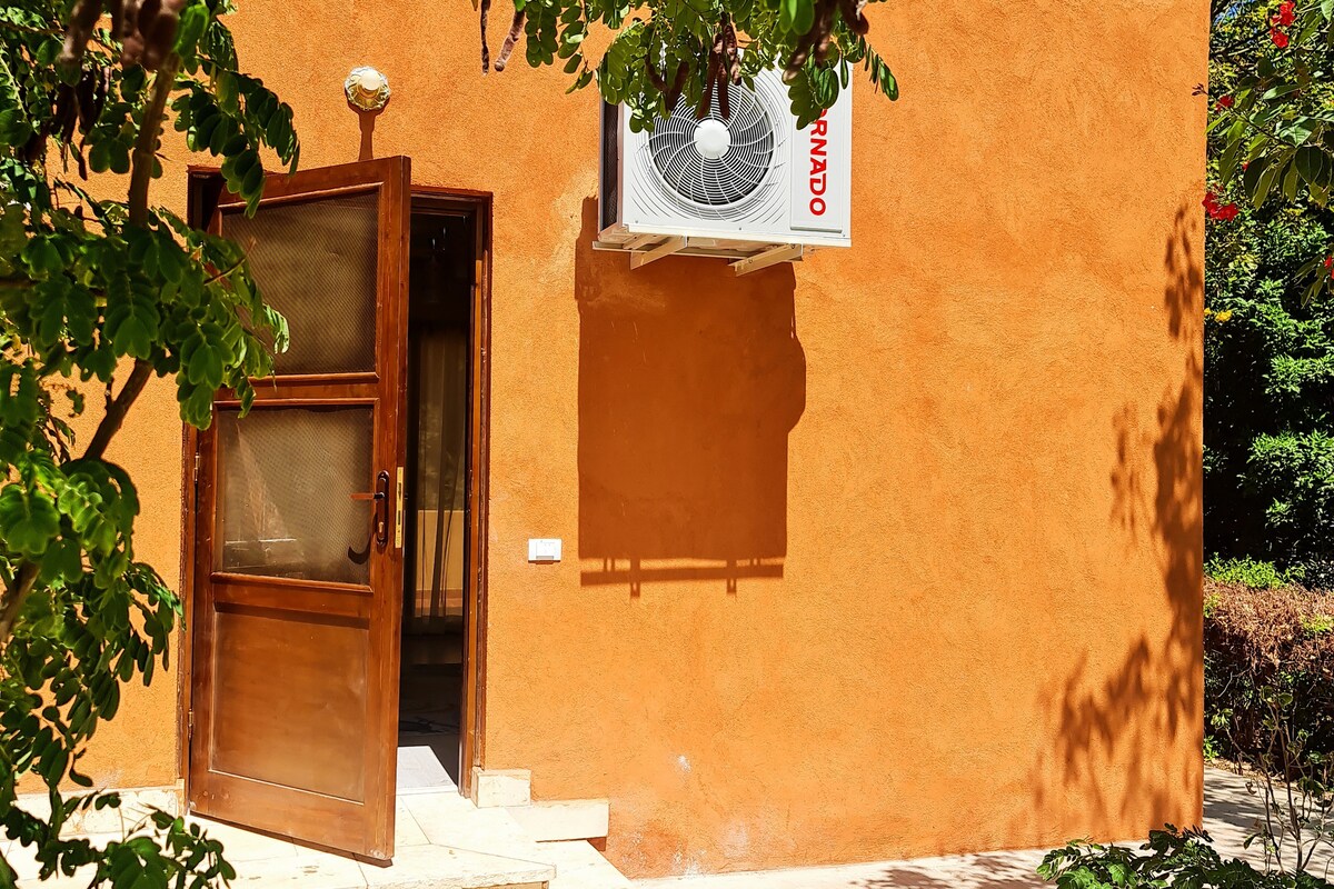 A wooden door is shown slightly ajar, leading into a separate cottage with an earthy orange exterior. An air conditioning unit is mounted on the wall beside the entrance. Lush greenery encircles the entrance, adding a natural touch to the exterior.