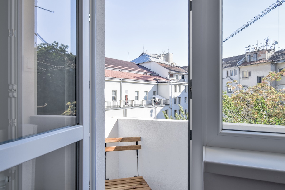 A small balcony is visible, featuring a wooden table and chair set. The view includes nearby buildings and clear blue skies, providing a refreshing outdoor space. Large windows frame the scene, allowing natural light to brighten the interior.