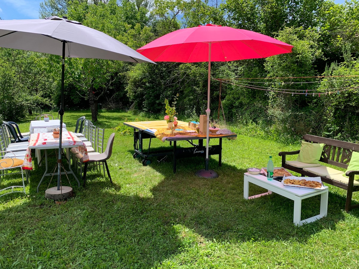 A spacious outdoor area is visible, featuring a wooden table set under a pink and grey umbrella. Nearby, a picnic table is adorned with a tablecloth and surrounded by various chairs. Lush greenery surrounds the space, creating a relaxed atmosphere.