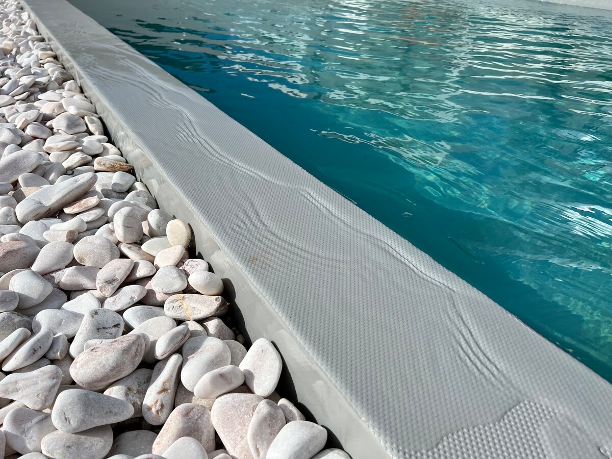 A close-up view of the edge of a private infinity pool reveals a textured surface, surrounded by smooth white pebbles. The clear blue water creates a calming reflection, enhancing the serene atmosphere of the outdoor space.