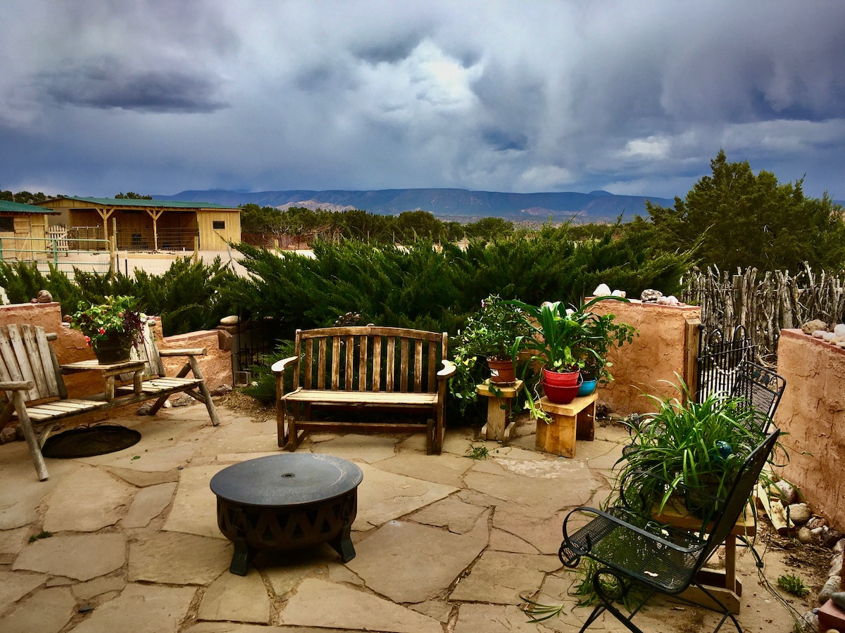 An outdoor patio features stone flooring with a rustic fire pit at its center. Surrounding seating includes wooden benches and a metal chair. Potted plants add greenery, and distant mountains are visible against a moody sky. A fence enhances the natural ambiance.