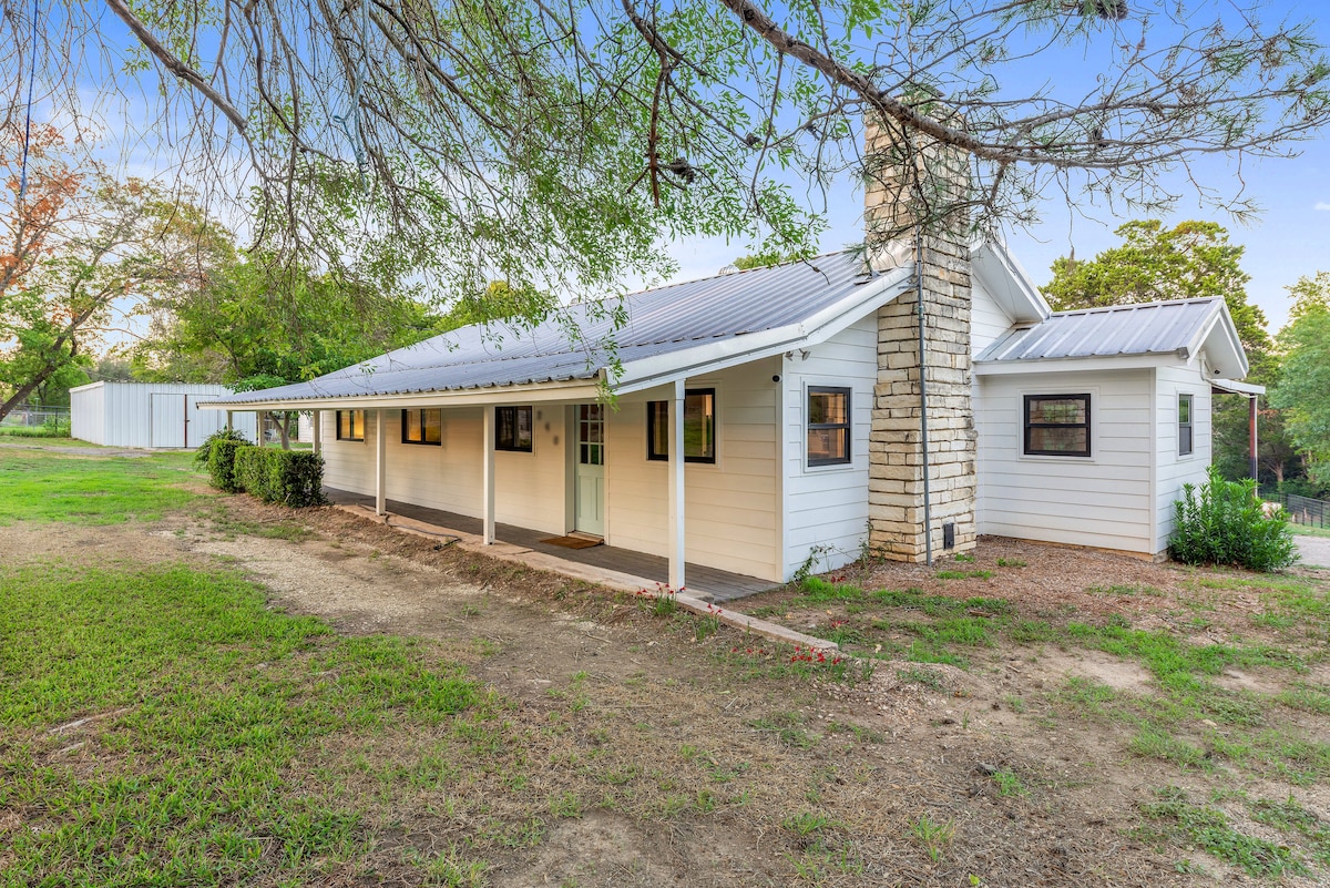 A single-story home is presented with a light exterior, featuring a stone chimney and overhanging roof. A covered porch extends along the front, bordered by green grass and trees, providing a tranquil outdoor space. The surrounding area features a well-maintained landscape.