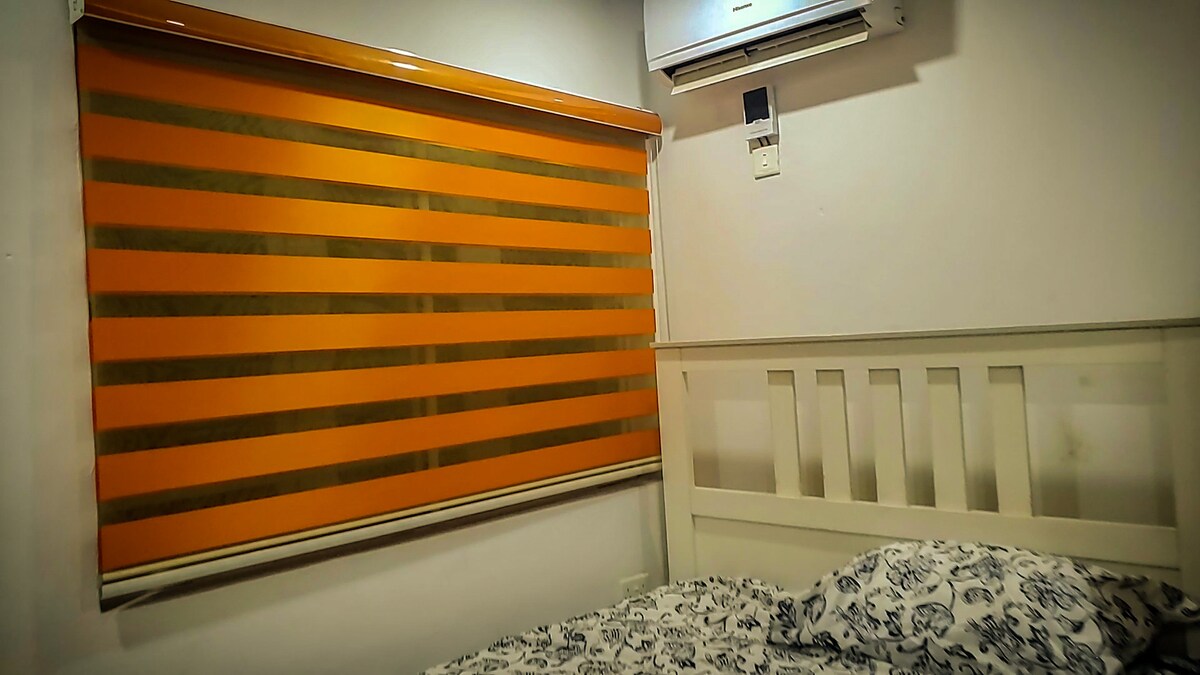 A cozy bedroom is presented with a white wooden bed adorned with patterned bedding. An orange and beige striped roller blind filters natural light, while an air conditioning unit is mounted above for climate control.
