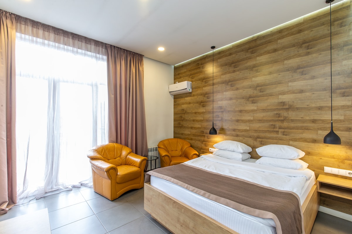 The studio features a spacious bed with white bedding arranged neatly. Two brown armchairs are positioned next to a large window with sheer curtains, allowing natural light to brighten the room. Soft ambient lighting is provided by pendant lights mounted on the wood-paneled wall.