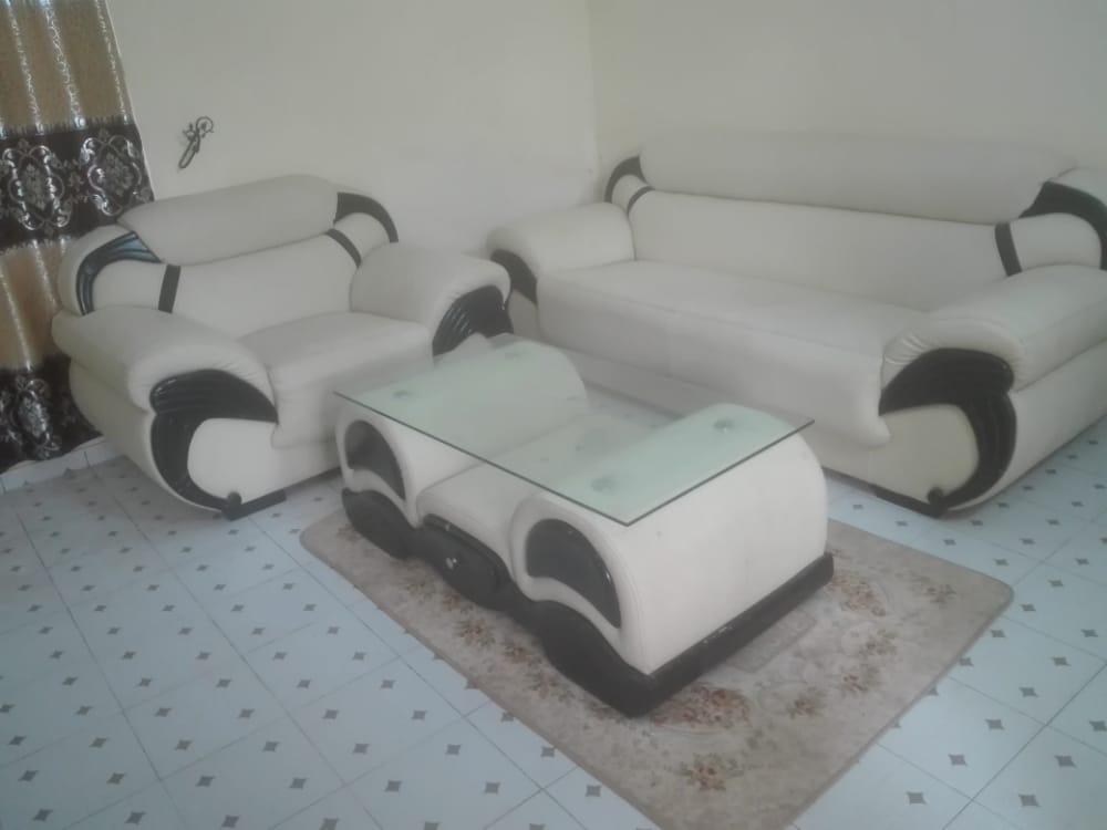 A light-colored living room features a white leather sofa set with contrasting black accents, including a spacious three-seater and a two-seater sofa. A glass coffee table sits atop a patterned area rug, and patterned curtains frame the window, adding texture to the space.